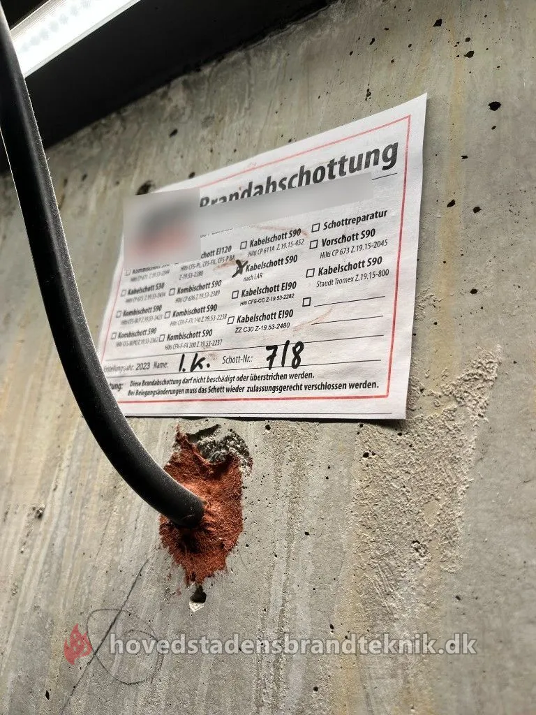 Kabel ført gennem betonvæg, tætnet med rødt brandtætningsmateriale og mærket med skilt